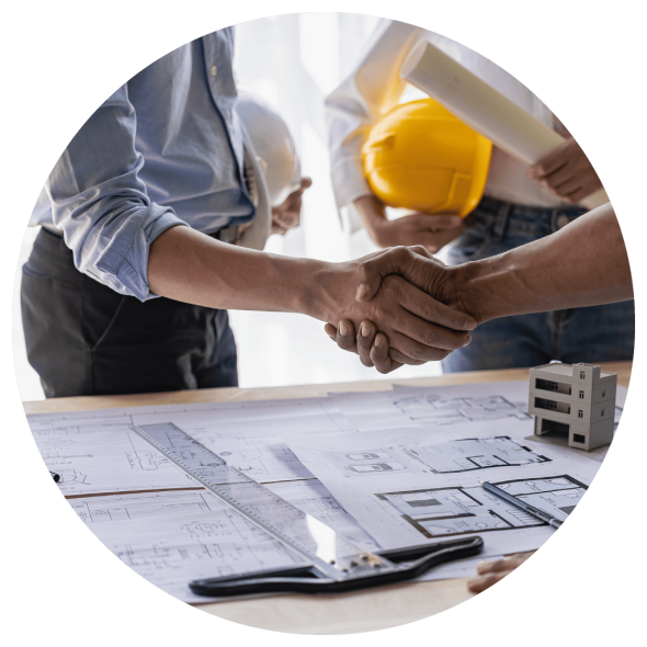 Three men holding hard hats are conversing over a desk with architectural plans and a model of a building; two of the men are shaking hands.